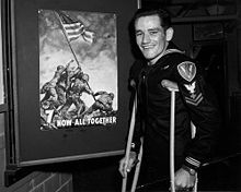 約翰·布拉得利，站在一幅向人們宣傳購買軍券的海報旁。當時他由於在一次戰鬥中被彈片擊中而受傷，暫時需要拐杖。